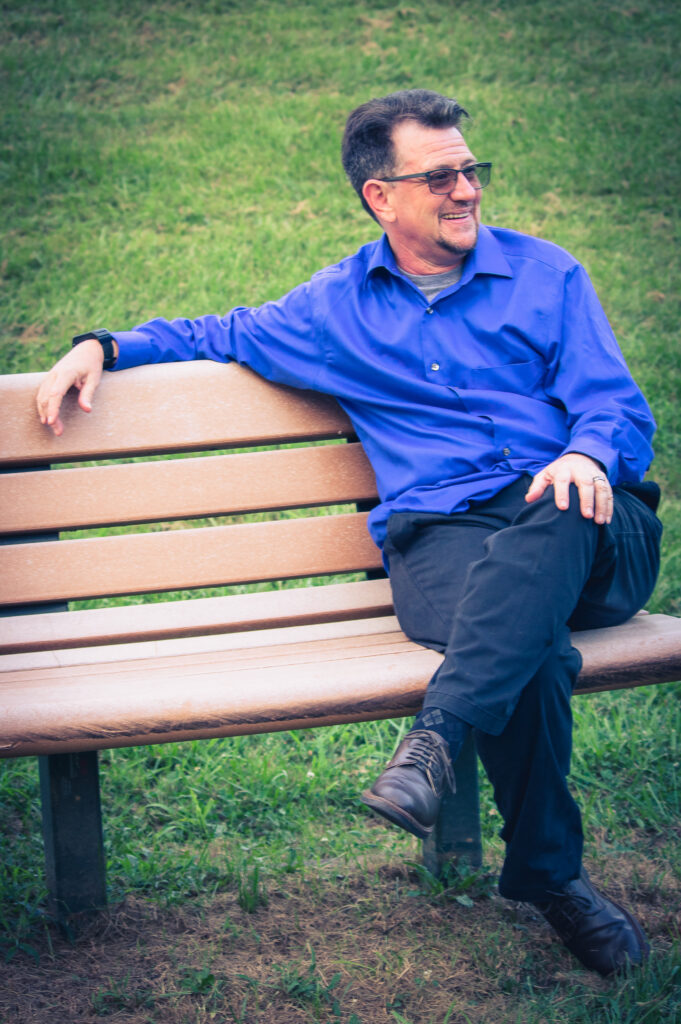 Don sitting on a park bench with one arm over the back of the bench. He is wearing a blue shirt. He is looking to his left and smiling.